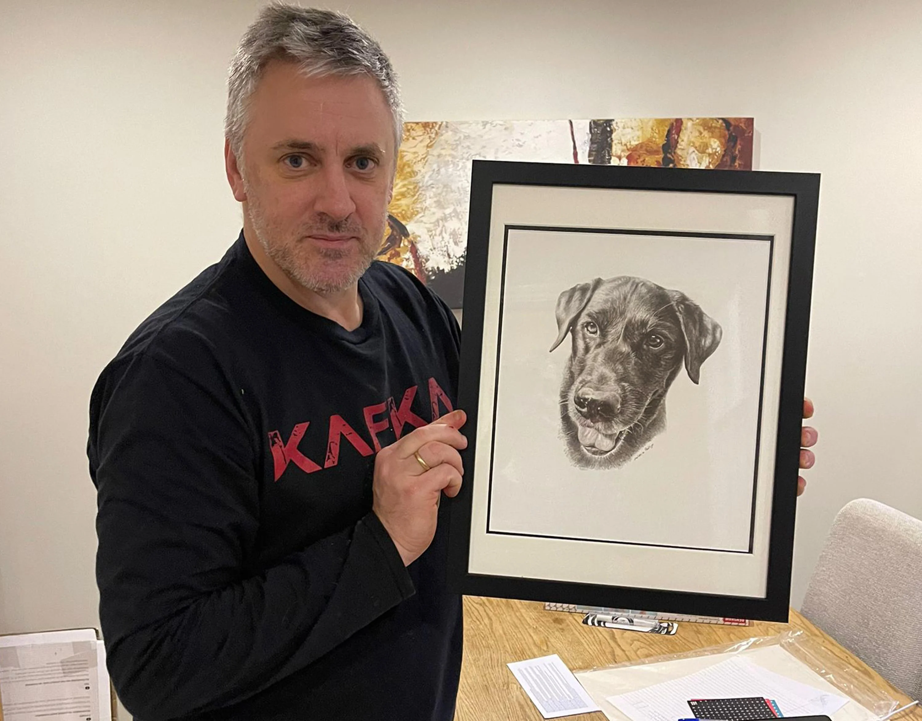 Framed pencil portrait of a black Labrador (Beano) being proudly held by client Adam, showing his happy reaction.