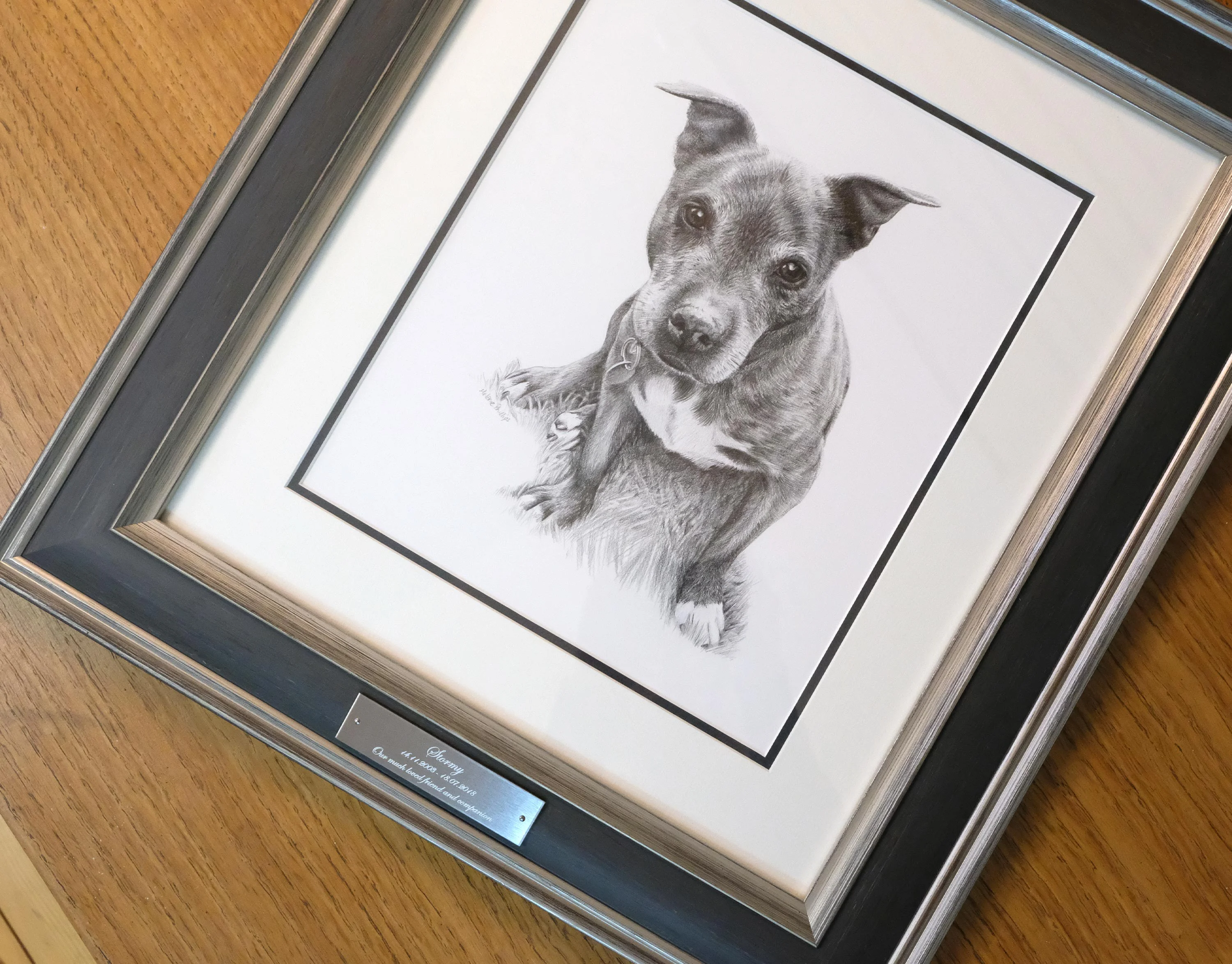 Framed pencil pet portrait of a Staffie dog displayed on a wooden table with an engraved nameplate.