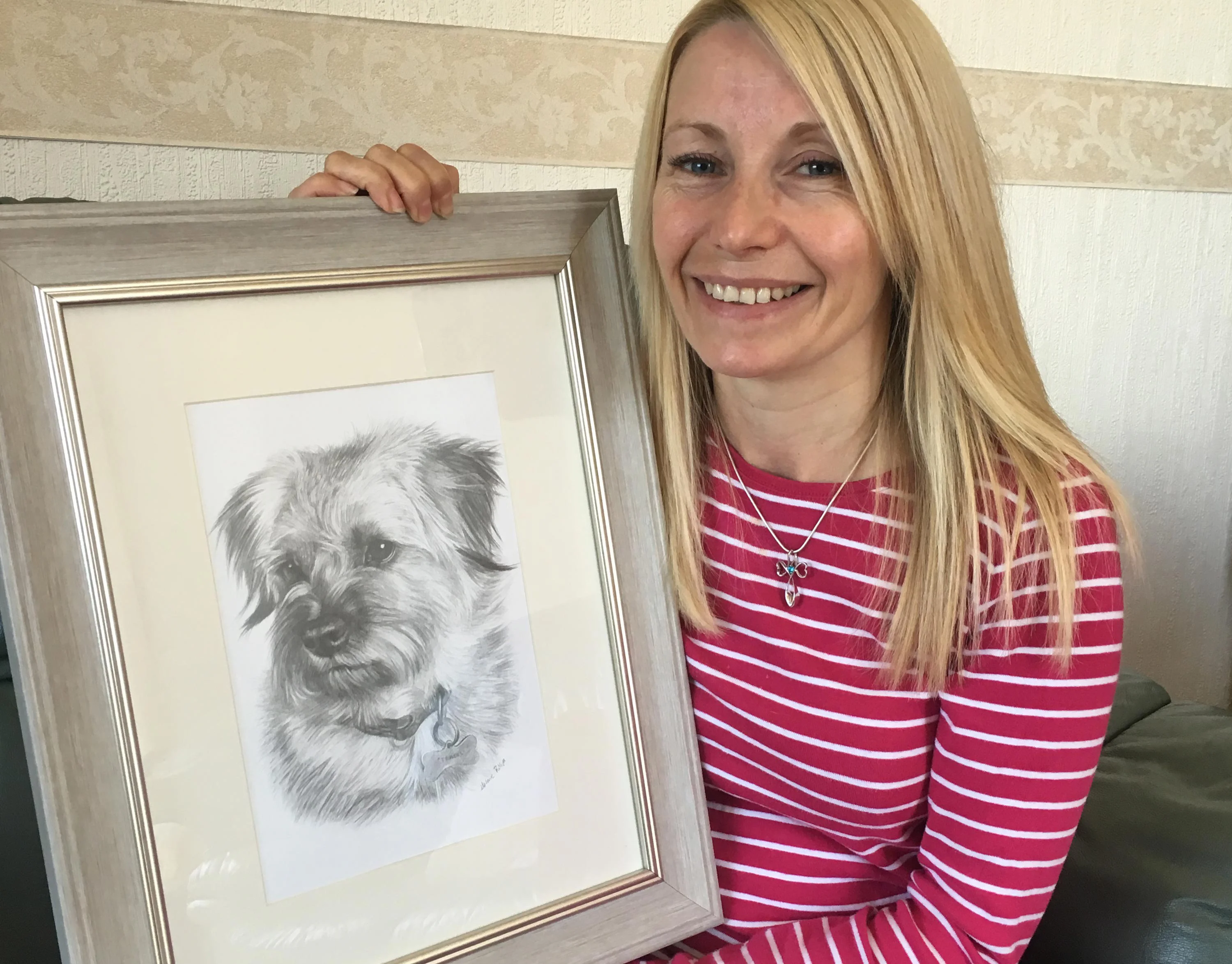 Happy client, Paula, holding a framed pencil portrait of her Border Terrier cross dog, Teasel.
