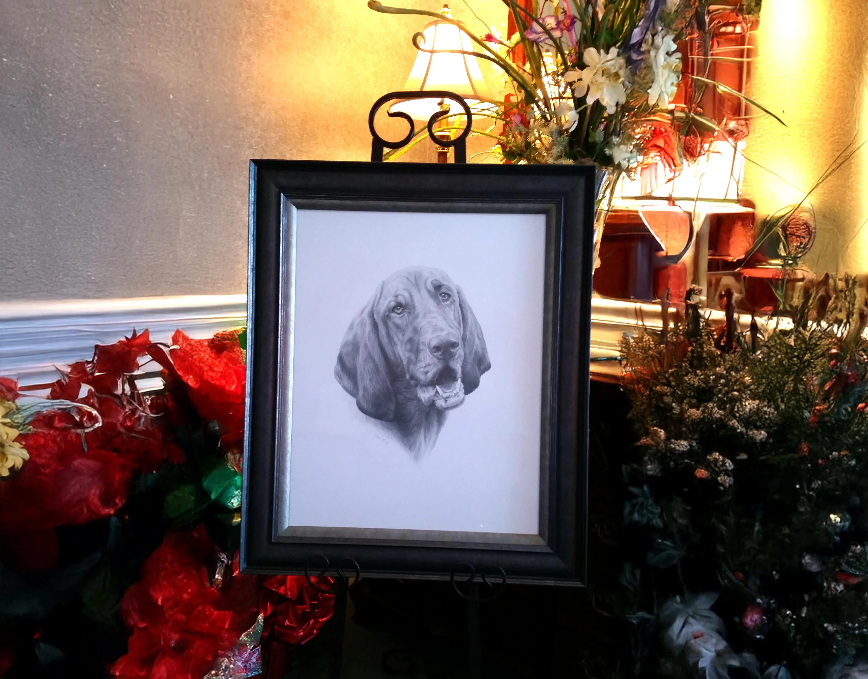 Framed pencil drawing of a Bloodhound (Daisy) on display, surrounded by decorative flowers and illuminated by a warm light.