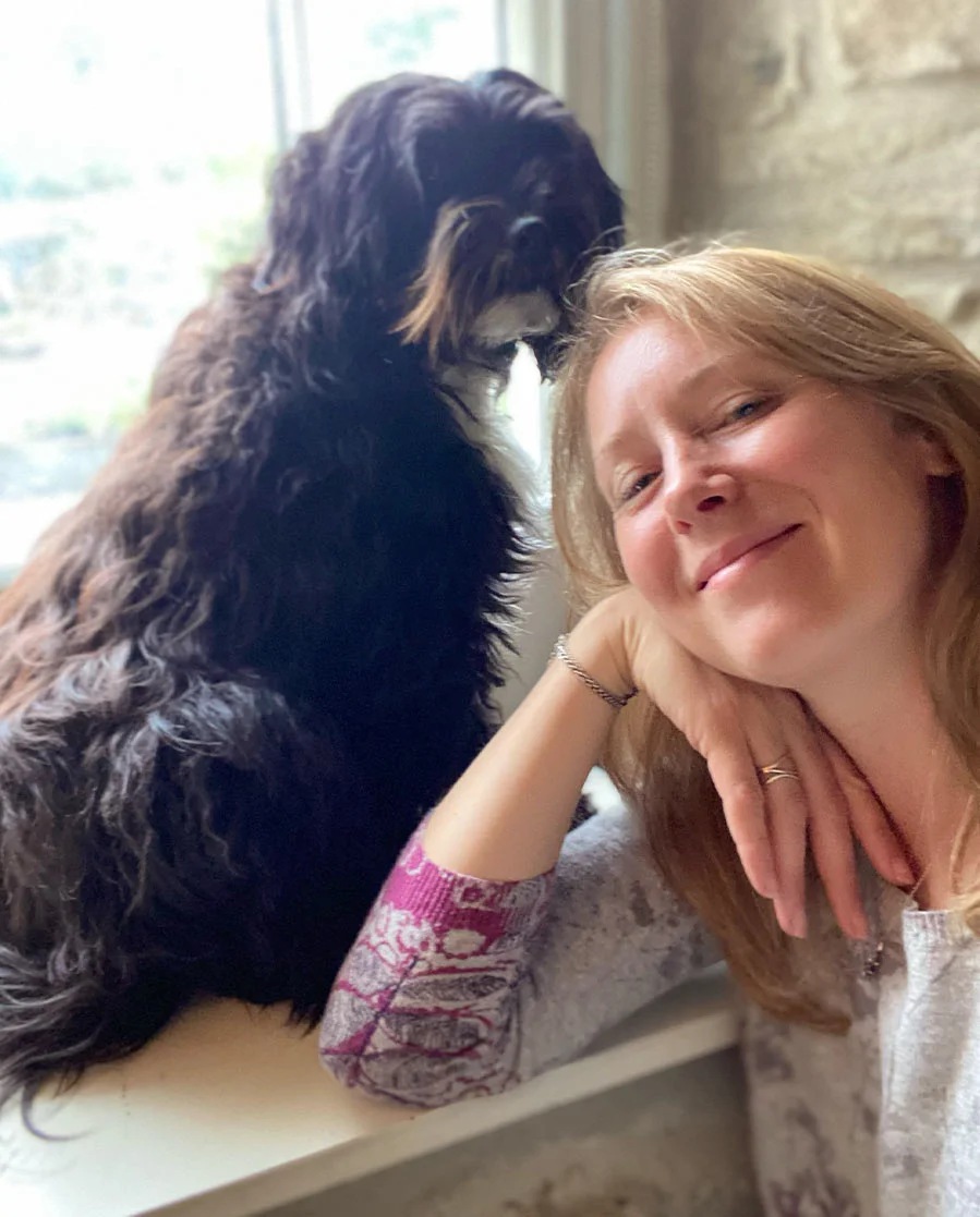Melanie Phillips smiling with her Tibetan Terrier, Lily, inside their cozy Welsh cottage.