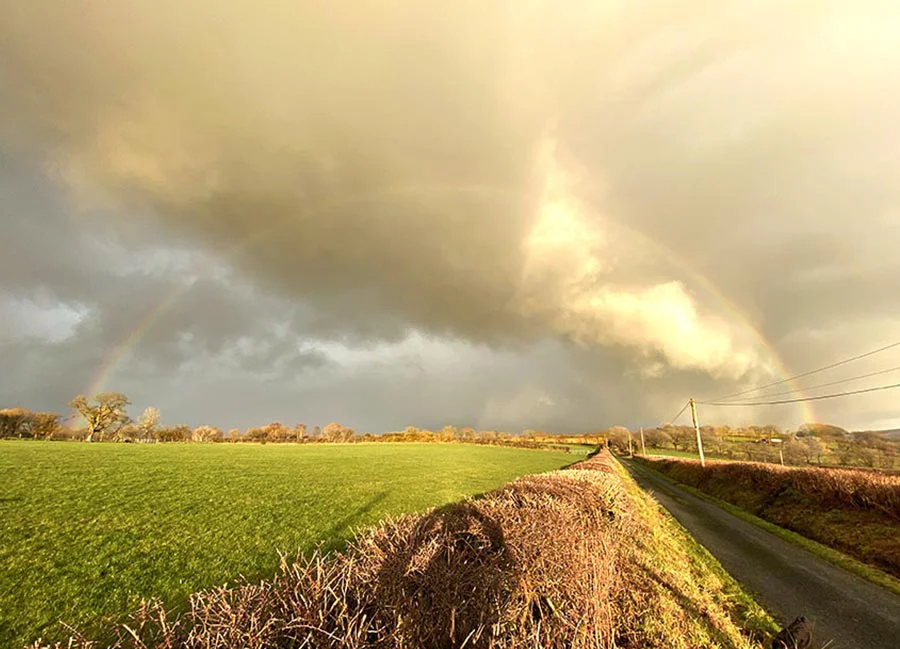 A photograph taken by Melanie Phillips showcasing dramatic clouds and a vibrant rainbow across the sky.
